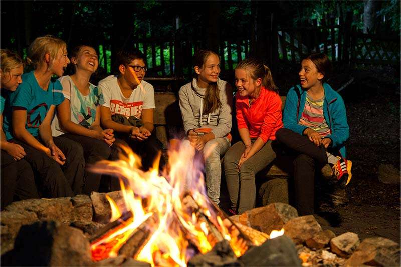 ThementagOutdoorKarte Eine Gruppe Jugendlicher sitzt am Abend gemeinsam um ein Lagerfeuer im Wald, lacht und unterhält sich.